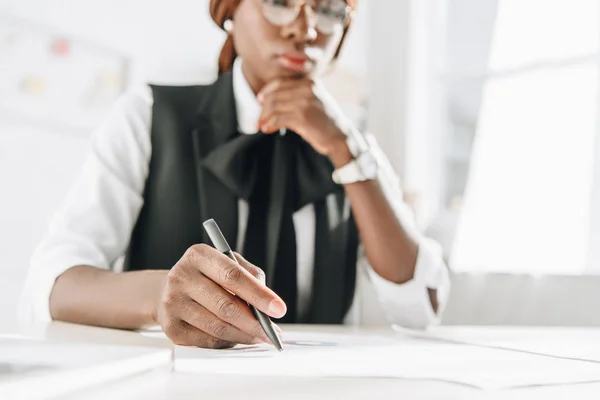 Focus sélectif de l'architecte afro-américaine adulte dans les lunettes en utilisant un stylo et en travaillant sur des documents au bureau — Photo de stock