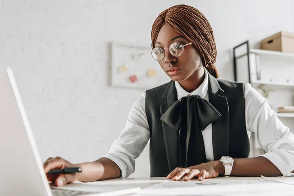 Attraente focalizzata afro-americana adulta donna d'affari in occhiali seduta alla scrivania del computer e al lavoro in ufficio — Foto stock