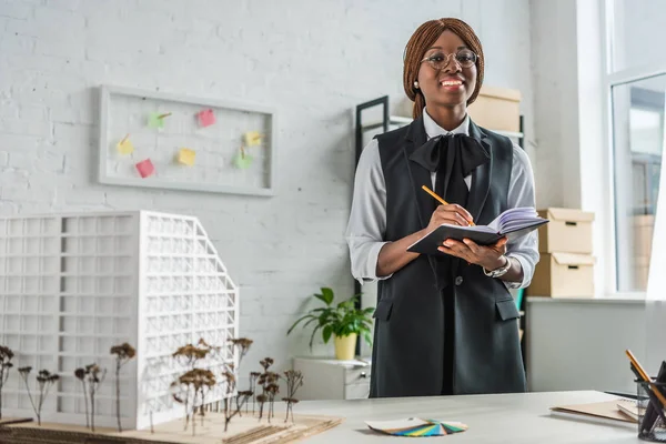 Sorridente afro-americana arquiteta adulta em óculos olhando para câmera escrevendo em notebook e trabalhando em projeto de construção no escritório — Fotografia de Stock