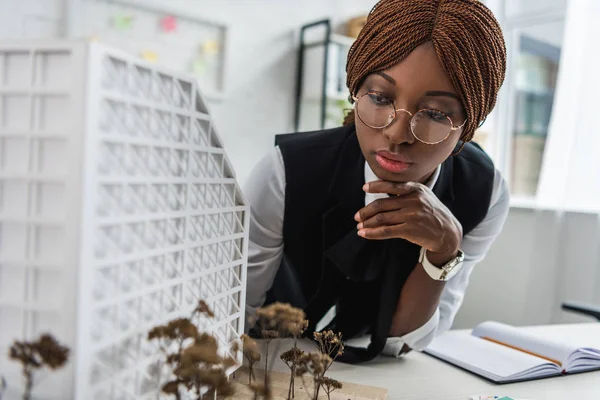 Serious african american female adult architect in glasses and formal wear touching chin with hand and working on construction project — Stock Photo