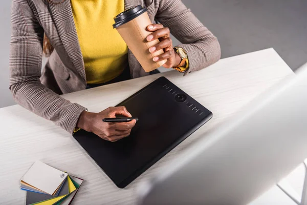 Teilansicht eines afrikanisch-amerikanischen Designers, der Coffee to go hält und am Arbeitsplatz ein Grafik-Tablet benutzt — Stockfoto