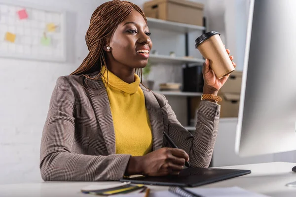 Sorridente adulto afro-americano adulto empresária sentada na mesa de computador, bebendo café e usando tablet gráfico — Fotografia de Stock