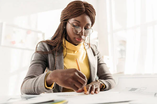 Focalizzata afro-americana architetto donna in bicchieri che tiene la matita e lavora al progetto alla scrivania con cianografie in ufficio — Foto stock