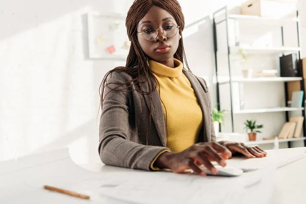 Arquitecta afroamericana concentrada en gafas y ropa formal trabajando en proyecto con planos en escritorio de oficina - foto de stock