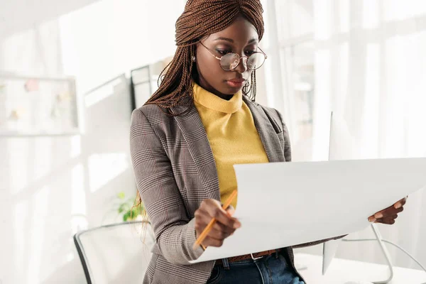 Femme architecte afro-américaine focalisée dans les lunettes tenant le plan et le crayon dans le bureau — Photo de stock