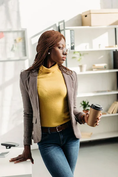 Bela mulher de negócios adulta afro-americana em desgaste formal e óculos segurando café para ir e posando no escritório moderno — Fotografia de Stock