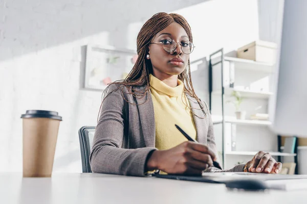 Konzentrierte afrikanisch-amerikanische Geschäftsfrau sitzt am Computer-Schreibtisch, bedient sich eines Grafik-Tablets und blickt am Arbeitsplatz in die Kamera — Stockfoto