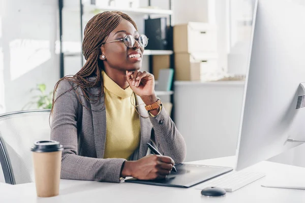 Улыбается африканский взрослый американский бизнесмен с помощью графического планшета и сидя за компьютерным столом в офисе — стоковое фото