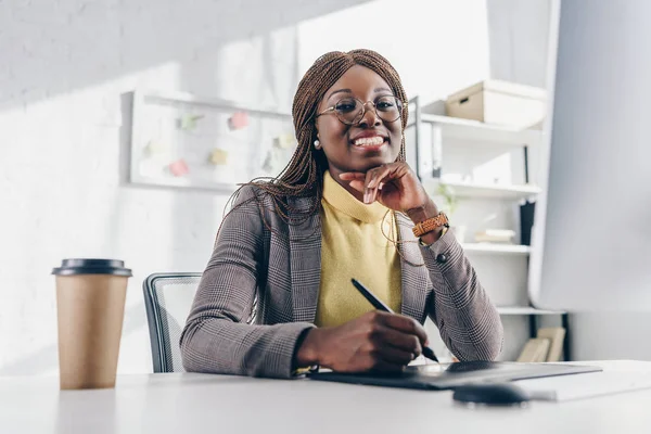 Lächelnde erwachsene afrikanisch-amerikanische Geschäftsfrau, die in die Kamera schaut, am Computertisch sitzt und ein Grafik-Tablet benutzt — Stockfoto