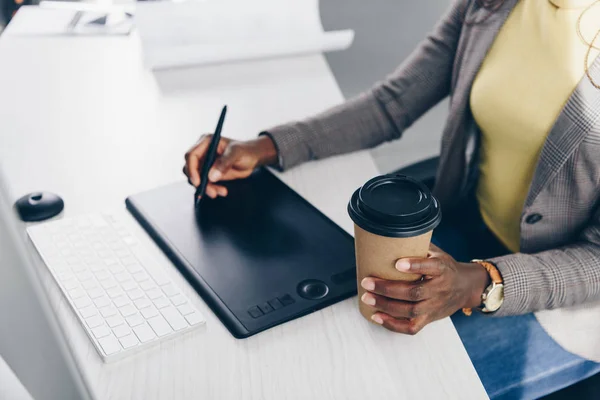 Teilansicht eines afrikanisch-amerikanischen Designers, der Coffee to go hält und am Arbeitsplatz ein Grafik-Tablet benutzt — Stockfoto