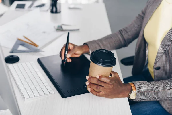 Vista parcial del diseñador afroamericano sosteniendo café para llevar y utilizando tableta gráfica en el lugar de trabajo - foto de stock