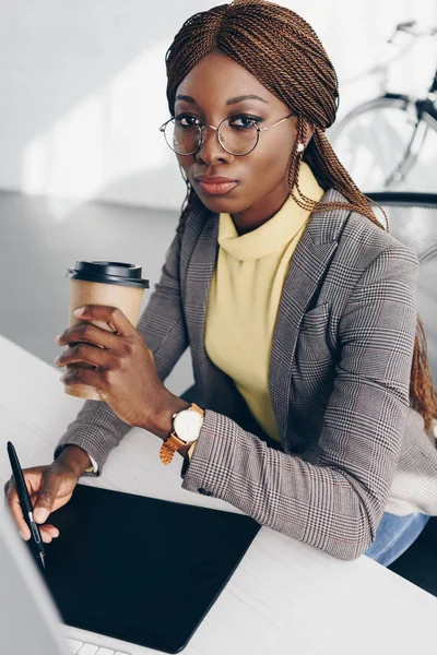 Belle femme d'affaires afro-américaine grave utilisant une tablette graphique, boire du café pour aller et regarder la caméra sur le lieu de travail — Photo de stock