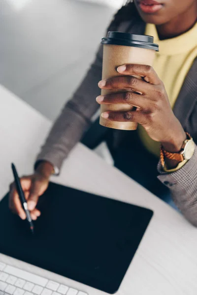 Vista parcial de la diseñadora afroamericana usando tableta gráfica y tomando café en el trabajo en la oficina - foto de stock
