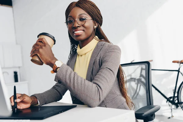 Schöne lächelnde afrikanisch-amerikanische Geschäftsfrau mit Grafik-Tablet, Kaffee to go trinken und am Arbeitsplatz in die Kamera schauen — Stockfoto