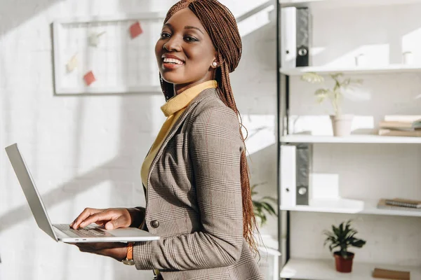 Africano americano adulto empresária segurando laptop no local de trabalho, sorrindo e olhando para a câmera — Fotografia de Stock