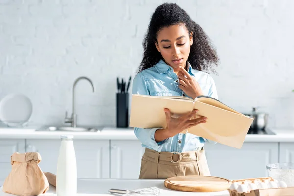 Стоїть приваблива афроамериканська дівчина дивиться на кулінарну книгу на кухні — стокове фото