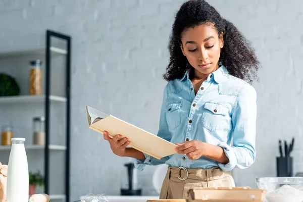Atractiva chica afroamericana sosteniendo libro de cocina en la cocina - foto de stock