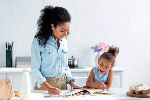 African american mother and daughter looking at cookbook in kitchen — Stock Photo