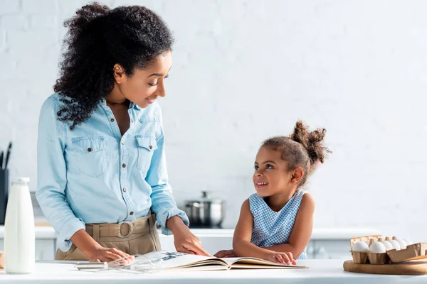 Cheerful african american mother and daughter looking at each other, cookbook on table in kitchen — Stock Photo