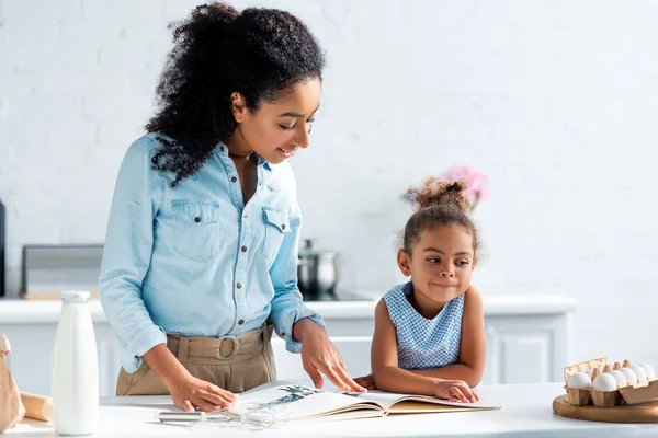 Madre e hija afroamericana leyendo libro de recetas en cocina - foto de stock