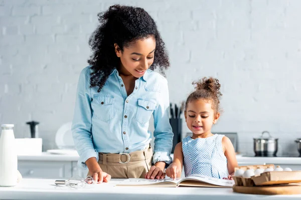Sorridente afro americano madre e figlia guardando libro di cucina insieme in cucina — Foto stock
