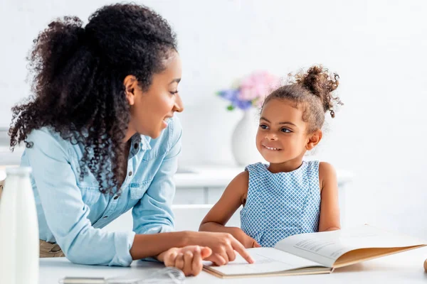 Sorrindo afro-americana mãe e filha olhando uns para os outros e escolhendo receita do livro de receitas na cozinha — Fotografia de Stock