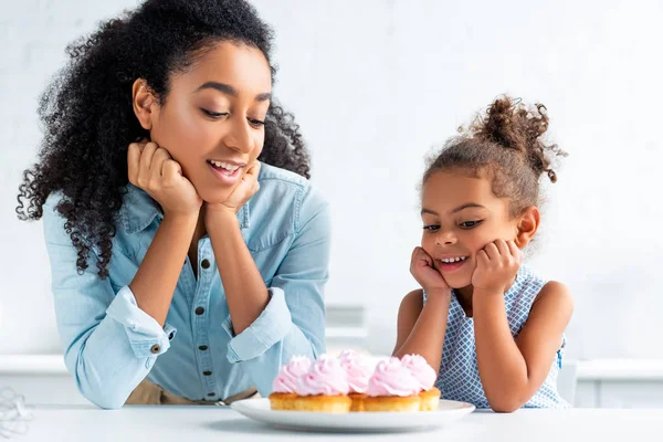 Cheerful african american mother and daughter resting chins on hands and looking at cupcakes on table in kitchen — Stock Photo