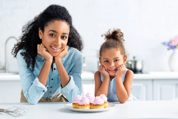 Alegre afroamericano madre e hija descansando barbilla en las manos y mirando a la cámara, cupcakes en la mesa en la cocina - foto de stock