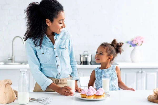 Madre e hija afroamericanas mirándose en la cocina, la leche y los cupcakes en la mesa - foto de stock