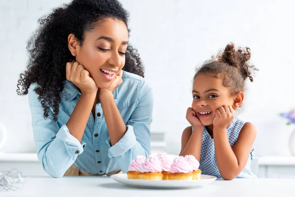 Happy african american mother and daughter resting chins on hands near cupcakes on table in kitchen — Stock Photo