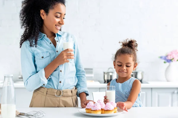 African american mother and daughter standing with glasses of milk and cupcakes in kitchen — Stock Photo