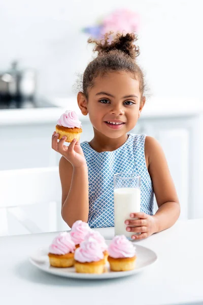 Adorable african american kid holding cupcake and glass of milk in kitchen, looking at camera — Stock Photo