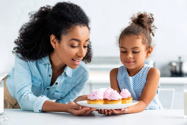 African american mother and daughter looking at homemade cupcakes in kitchen — Stock Photo