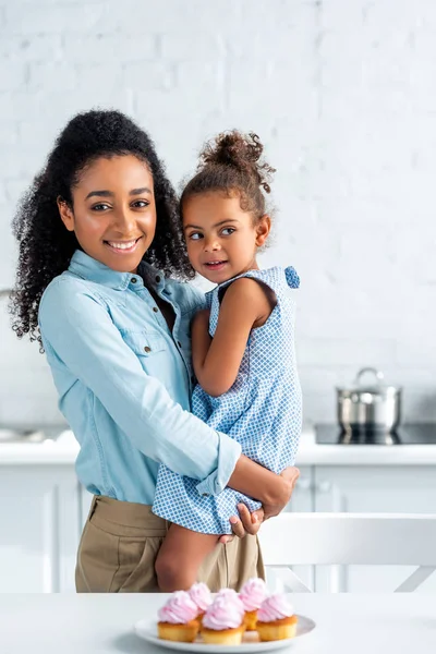 Glücklich afrikanisch-amerikanische Mutter umarmt Tochter in der Küche, Cupcakes auf dem Tisch — Stockfoto