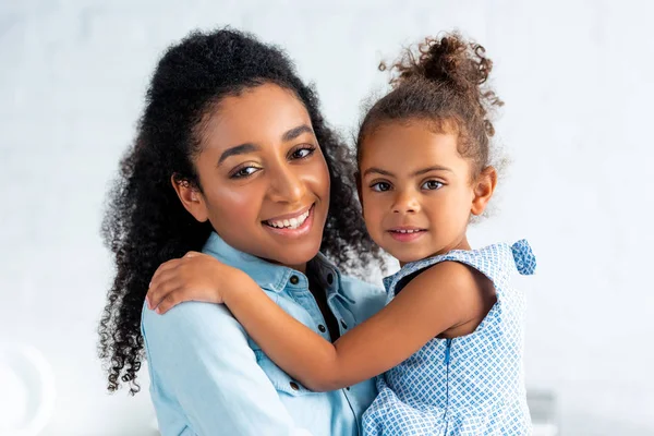 Sorridente afro-americano mãe e filha abraçando na cozinha e olhando para a câmera — Fotografia de Stock