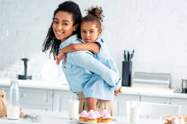 African american mother giving piggyback to daughter in kitchen, homemade cupcakes on table — Stock Photo