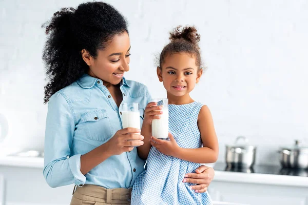 Sonriente afroamericana madre e hija sosteniendo vasos de leche en la cocina - foto de stock