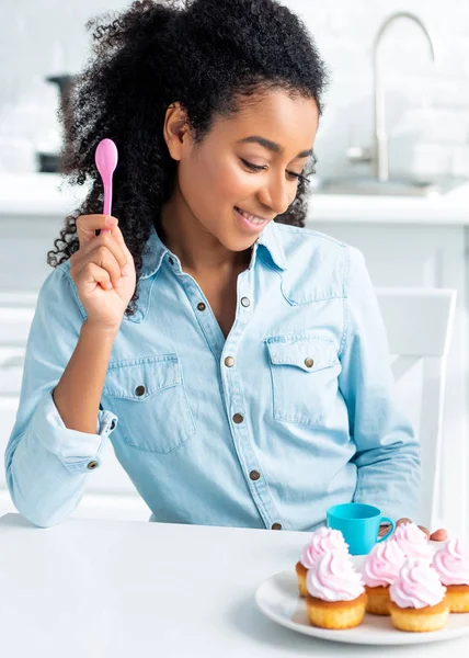 Sorrindo atraente menina afro-americana segurando colher de plástico e olhando para cupcakes na cozinha — Fotografia de Stock