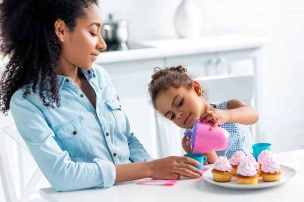 African american daughter and mother playing with plastic dishes in kitchen, kid pouring tea — Stock Photo