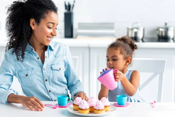 Улыбающаяся африканская американская дочь и мать играют с пластиковой посудой на кухне, ребенок наливает чай — стоковое фото