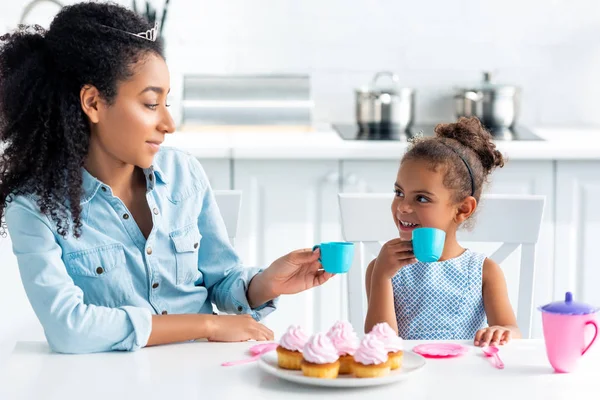 Afro-americana mãe e filha com tiaras segurando copos de plástico na cozinha — Fotografia de Stock