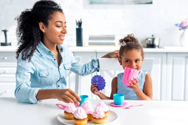 Alegre africano americano madre y hija con tiaras sentado a mesa con cupcakes en cocina - foto de stock