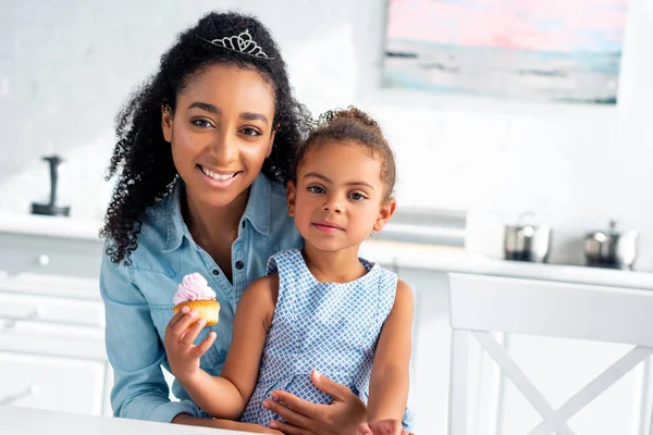 African american mother hugging daughter with cupcake in kitchen, looking at camera — Stock Photo