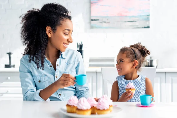 Sonriente afroamericana madre e hija con tiaras sosteniendo taza de plástico y magdalena en la cocina - foto de stock