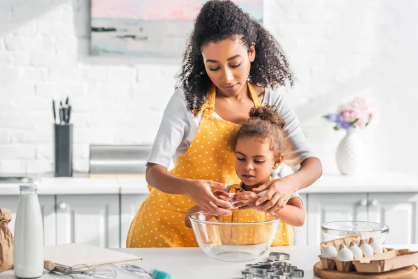 Madre afroamericana ayudando a su hija a romper huevo para preparar masa en la cocina - foto de stock