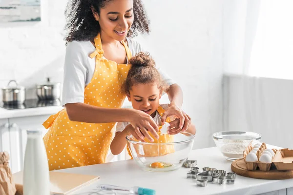 Smiling african american mother helping daughter breaking egg for preparing dessert in kitchen — Stock Photo