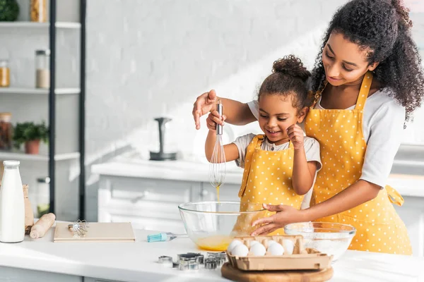 African american mother and daughter whisking eggs for dough in kitchen — Stock Photo