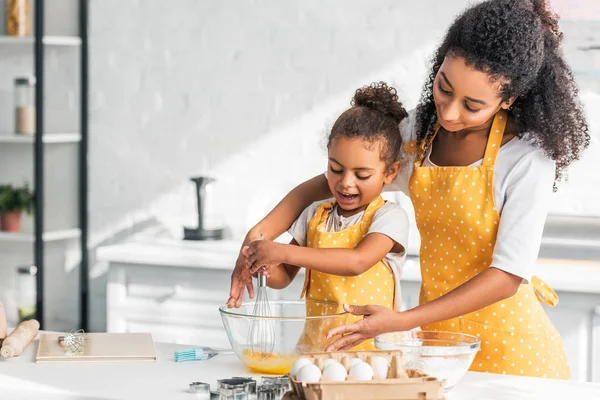 Mãe e filha africana americana em aventais amarelos batendo ovos para massa na cozinha — Fotografia de Stock