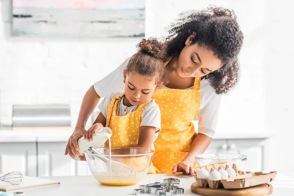 Afro-americana mãe e filha preparando massa e derramando leite em tigela na cozinha — Fotografia de Stock