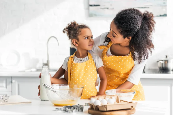 Madre e hija afroamericana mirándose durante la preparación de la masa en la cocina - foto de stock